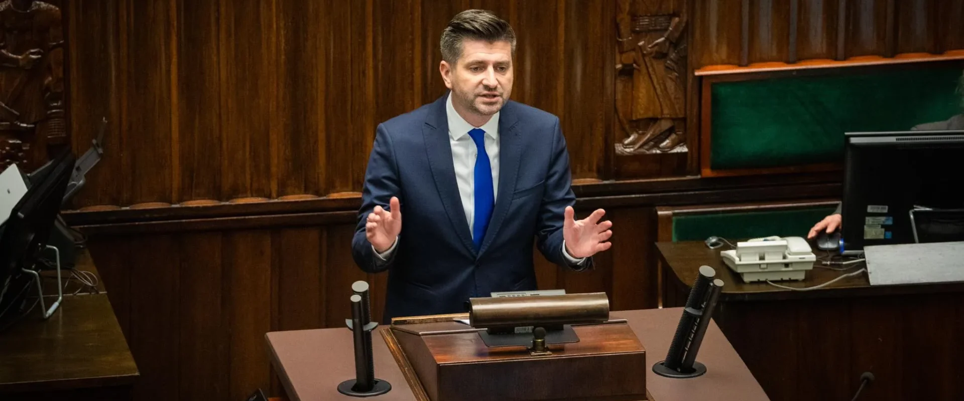 Mężczyzna w garniturze przemawia z mównicy w sali obrad, za nim widoczne drewniane panele i sprzęt biurowy.