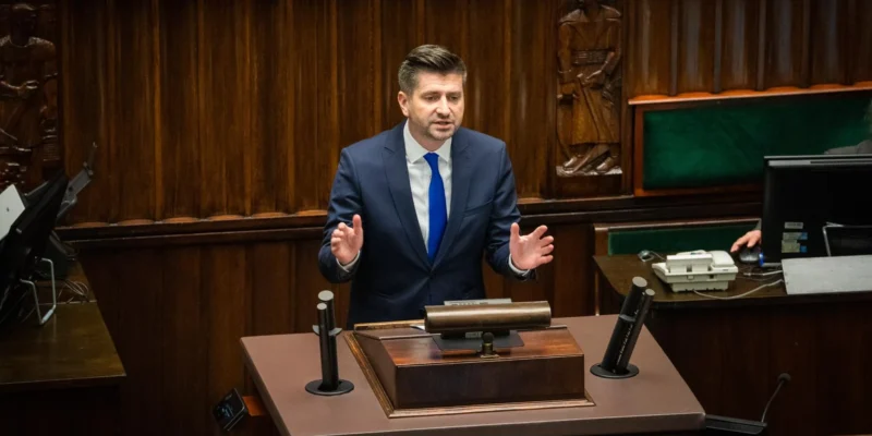 Mężczyzna w garniturze przemawia z mównicy w sali obrad, za nim widoczne drewniane panele i sprzęt biurowy.