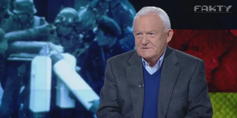 Starszy mężczyzna w marynarce i niebieskim swetrze siedzi w studiu telewizyjnym przed dużym ekranem z rozmytą grafiką i napisem "FAKTY".