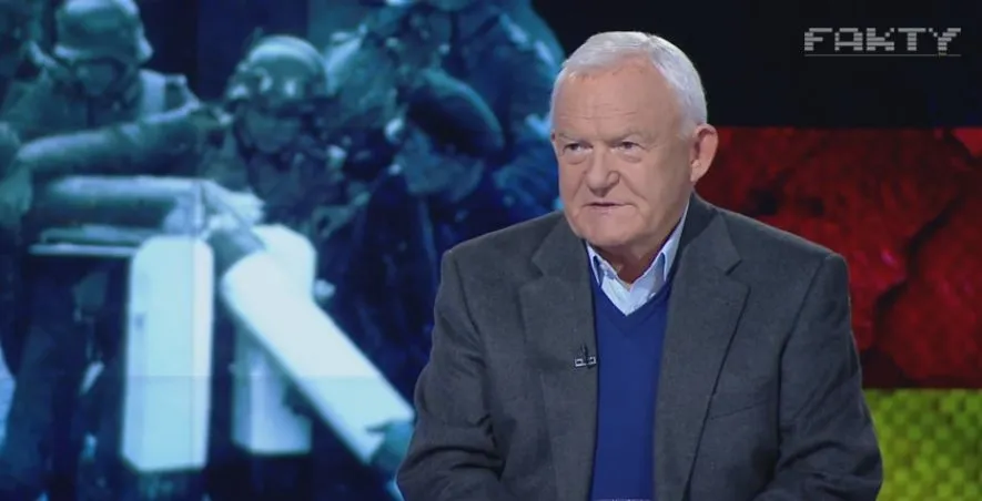 Starszy mężczyzna w marynarce i niebieskim swetrze siedzi w studiu telewizyjnym przed dużym ekranem z rozmytą grafiką i napisem "FAKTY".