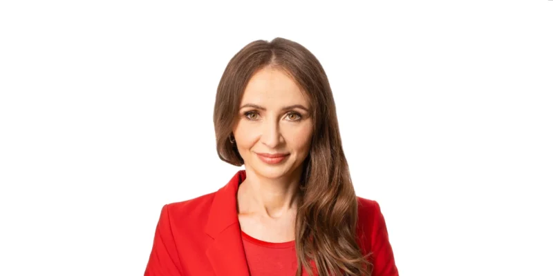 Kobieta w czerwonej marynarce i bluzce, z długimi włosami, uśmiecha się do kamery na tle białej ściany.