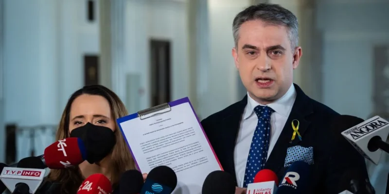 Mężczyzna w garniturze z żółtą wstążką na klapie trzyma na klipboardzie dokument podczas konferencji prasowej, otoczony przez liczne mikrofony mediów; obok niego stoi kobieta w maseczce.