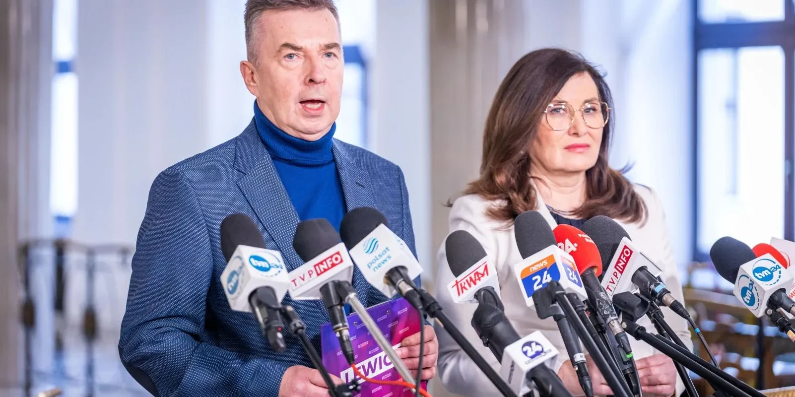 Na zdjęciu dwóch polityków stoi przed grupą mikrofonów, przygotowując się do konferencji prasowej.