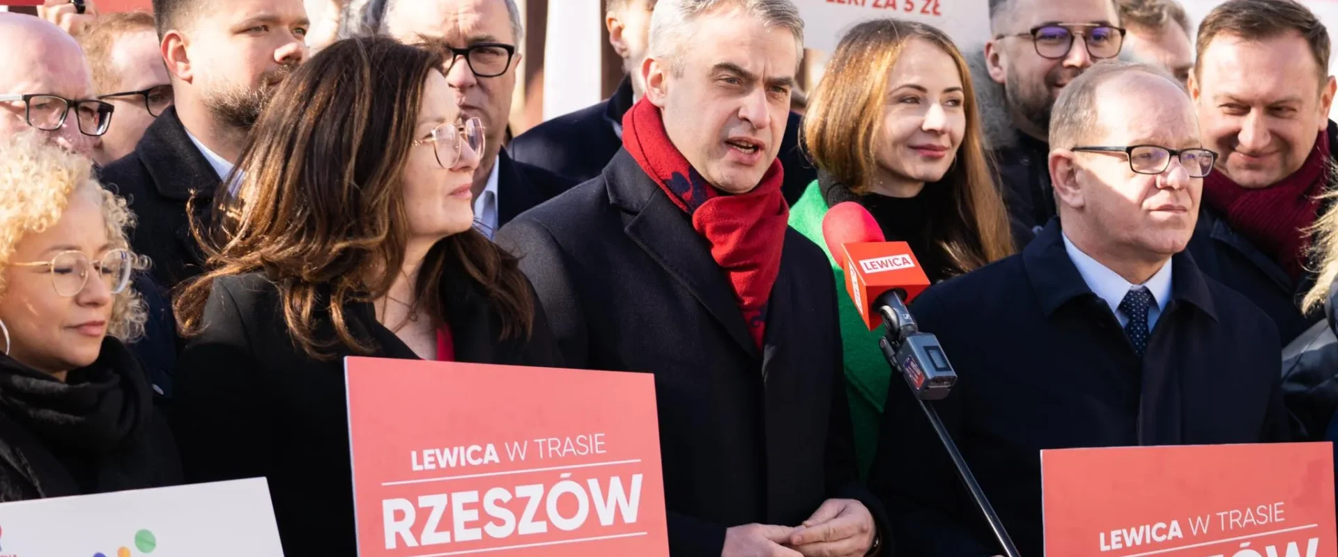 Na zdjęciu widoczna jest grupa ludzi na wiecu politycznym, trzymających czerwone plakaty z hasłem "Lewica w trasie Rzeszów".