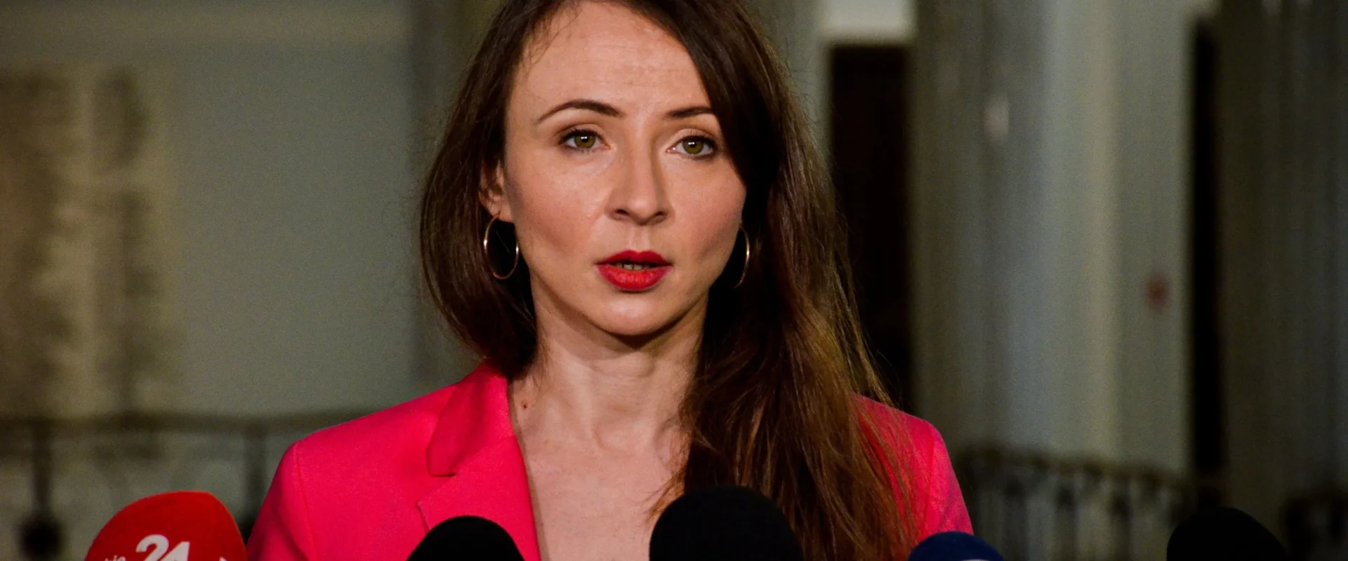 Kobieta w różowej marynarce przemawia na konferencji prasowej, stojąc przed licznymi mikrofonami z logo mediów.