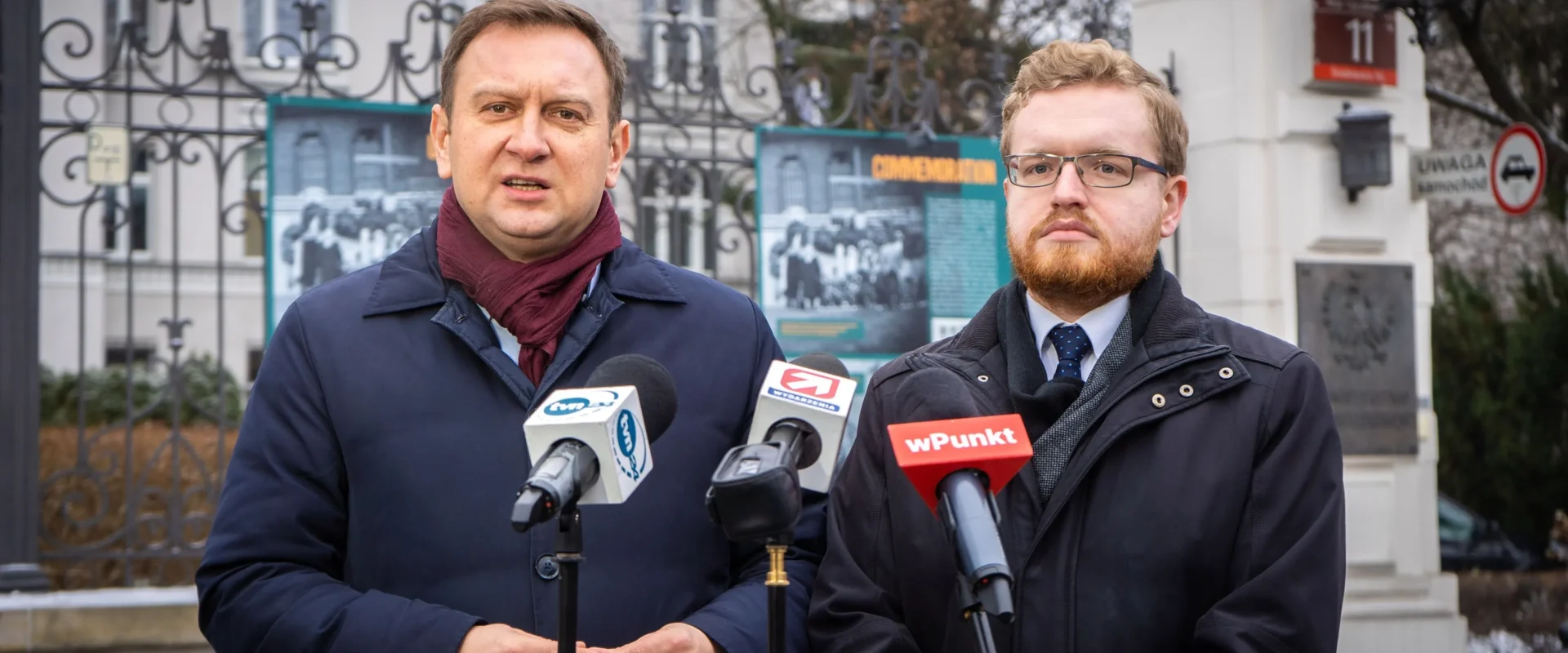Na zdjęciu prezentują się dwaj mężczyźni, którzy stoją przed mikrofonami podczas konferencji prasowej w otoczeniu budynków i plakatów informacyjnych.