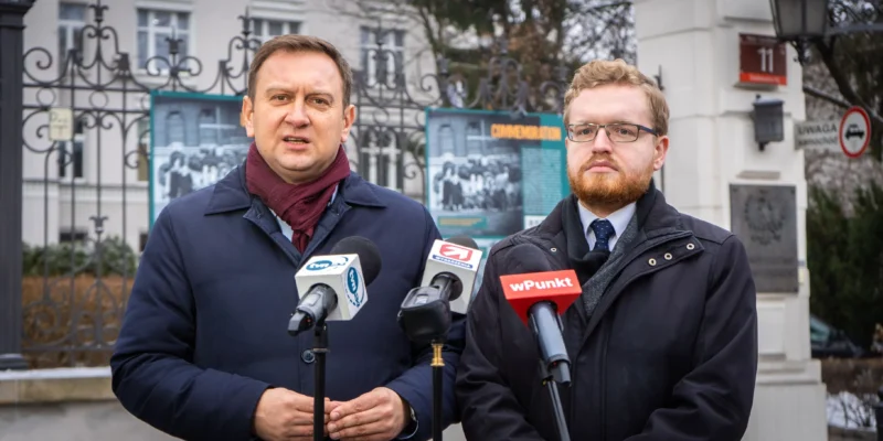 Na zdjęciu prezentują się dwaj mężczyźni, którzy stoją przed mikrofonami podczas konferencji prasowej w otoczeniu budynków i plakatów informacyjnych.