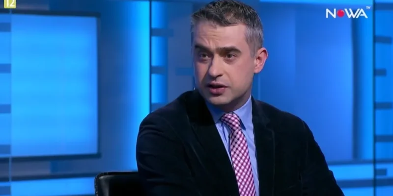 Mężczyzna w marynarce i różowo-białym krawacie siedzi i przemawia w niebiesko oświetlonym studiu telewizyjnym z widocznym logo NOWA TV.