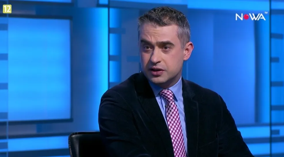 Mężczyzna w marynarce i różowo-białym krawacie siedzi i przemawia w niebiesko oświetlonym studiu telewizyjnym z widocznym logo NOWA TV.