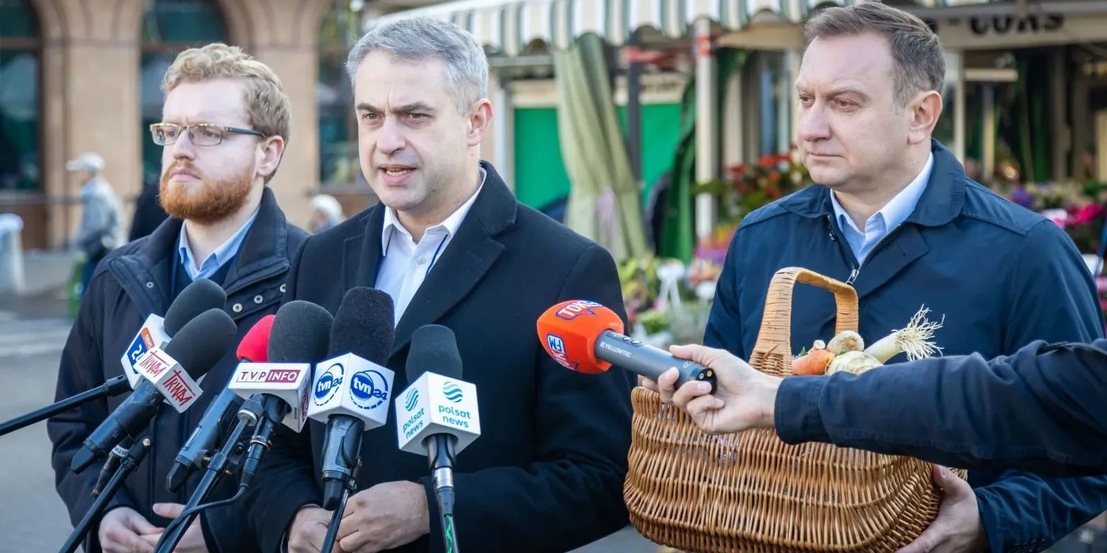 Trzech mężczyzn w garniturach przemawia na konferencji prasowej w otoczeniu kwiatów i warzyw, trzymając mikrofony oraz kosz z produktami.