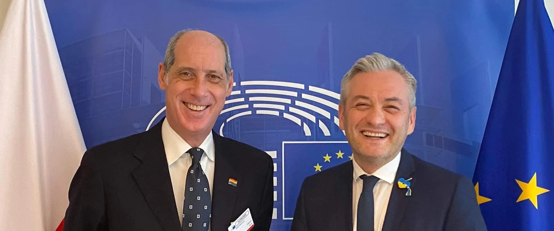 Dwóch mężczyzn w garniturach na tle flag Polski i Unii Europejskiej podczas oficjalnego spotkania.