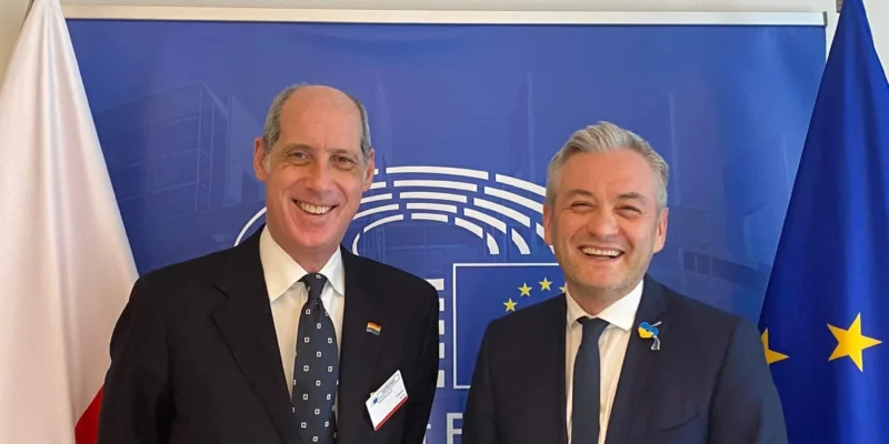 Dwóch mężczyzn w garniturach na tle flag Polski i Unii Europejskiej podczas oficjalnego spotkania.