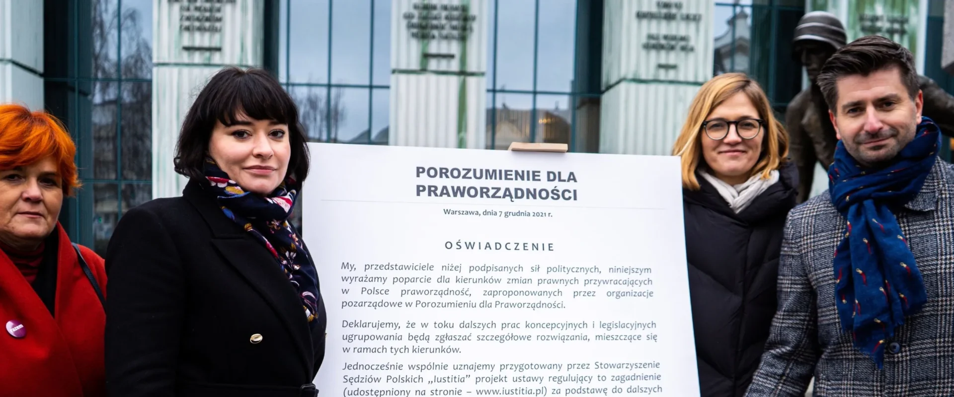 Na zdjęciu przedstawione są cztery osoby pozujące przed tablicą z oświadczeniem dotyczącym poparcia dla praworządności w Polsce.