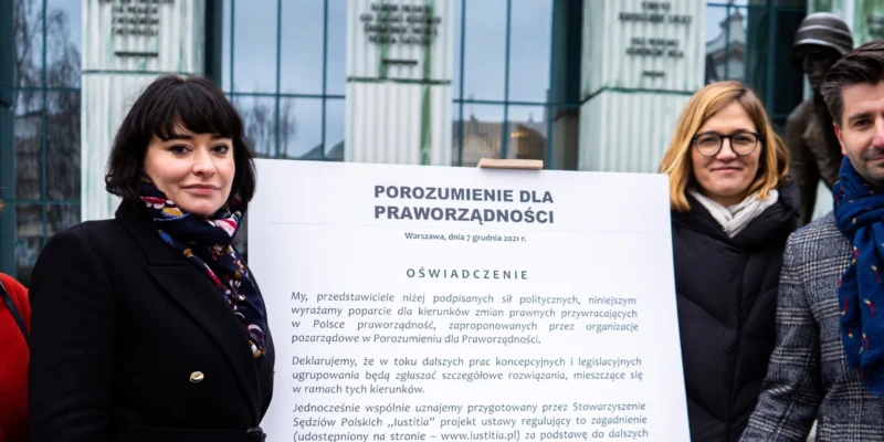 Na zdjęciu przedstawione są cztery osoby pozujące przed tablicą z oświadczeniem dotyczącym poparcia dla praworządności w Polsce.