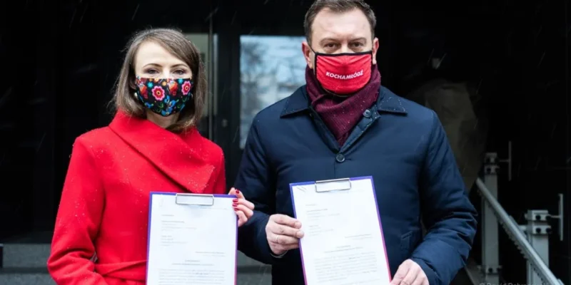 Dwoje osób w maseczkach trzyma pliki z dokumentami na tle wejścia do budynku.