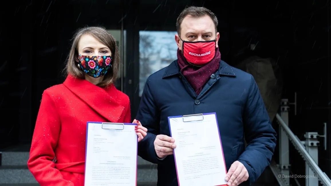 Dwoje osób w maseczkach trzyma pliki z dokumentami na tle wejścia do budynku.
