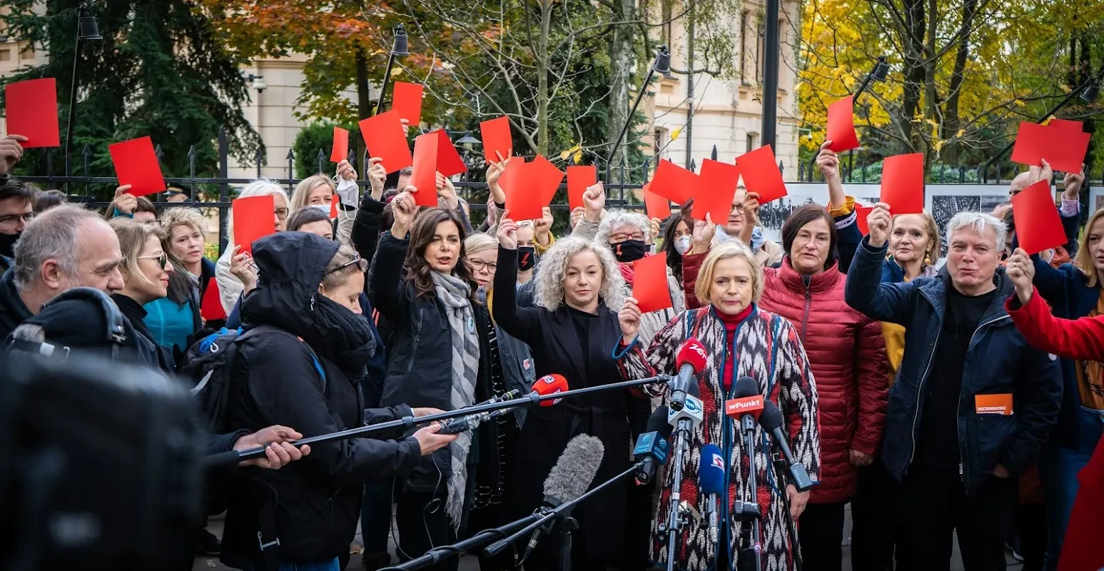 Grupa ludzi na demonstracji trzyma czerwone kartki nad głowami przed licznymi mikrofonami i kamerami w rocznicę wyroku wobec kobiet.
