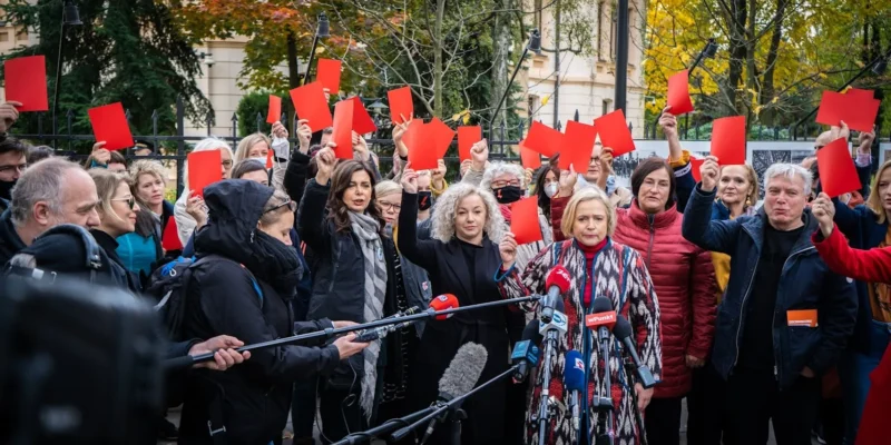 Grupa ludzi na demonstracji trzyma czerwone kartki nad głowami przed licznymi mikrofonami i kamerami w rocznicę wyroku wobec kobiet.