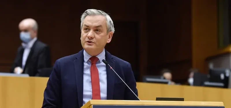 Polityk w granatowym garniturze ze czerwonym krawatem przemawia z mównicy w sali plenarnej, a w tle widoczni są urzędnicy w maseczkach.