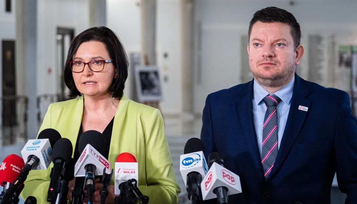 Na zdjęciu widoczna jest dwójka mówiących przy mikrofonach w budynku, najprawdopodobniej podczas konferencji prasowej.