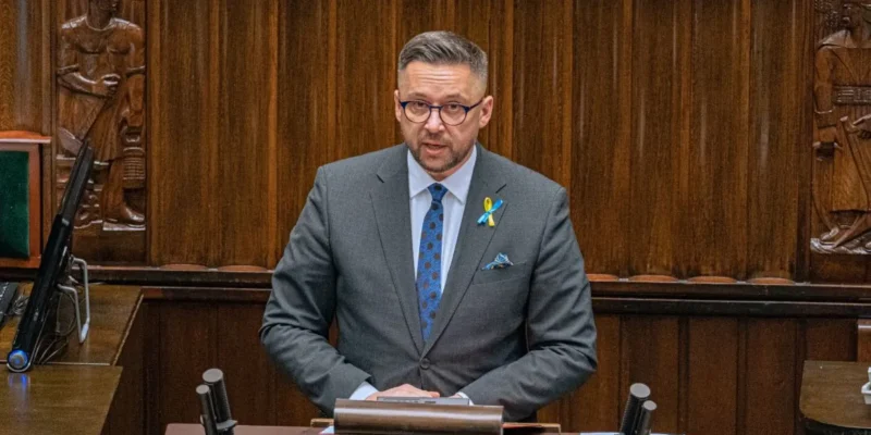 Mężczyzna w eleganckim garniturze przemawia w parlamencie, stojąc za mównicą na tle drewnianej ściany z rzeźbami.