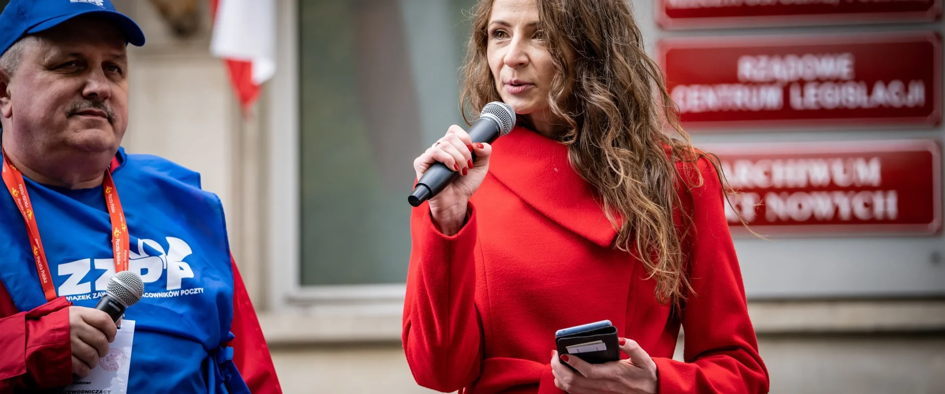 Kobieta w czerwonym płaszczu przemawia do mikrofonu podczas wydarzenia, obok niej stoi mężczyzna w niebieskiej koszulce z logo ZZPP.