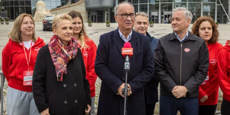 Grupa polityków Lewicy stoi na placu przed nowoczesnym budynkiem, a centralnie mężczyzna w garniturze przemawia do kamery z czerwonym mikrofonem z logo Lewica.