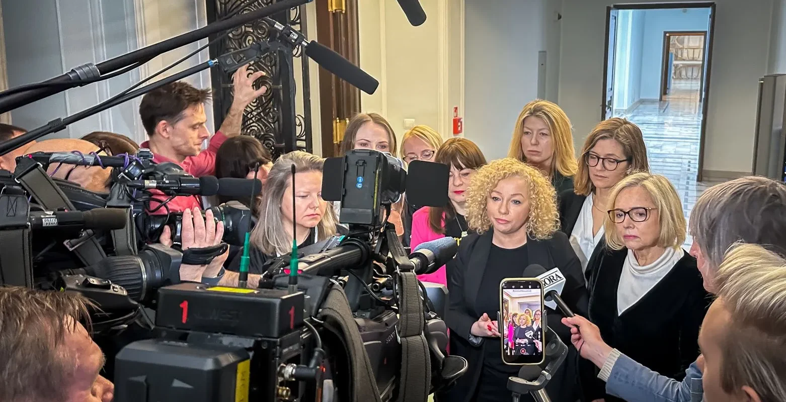 Grupa kobiet i mężczyzn w profesjonalnym otoczeniu medialnym, z kamerami i mikrofonami, stojąca przed mównicą w trakcie konferencji prasowej.