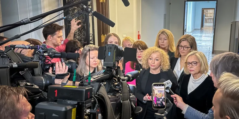 Grupa kobiet i mężczyzn w profesjonalnym otoczeniu medialnym, z kamerami i mikrofonami, stojąca przed mównicą w trakcie konferencji prasowej.