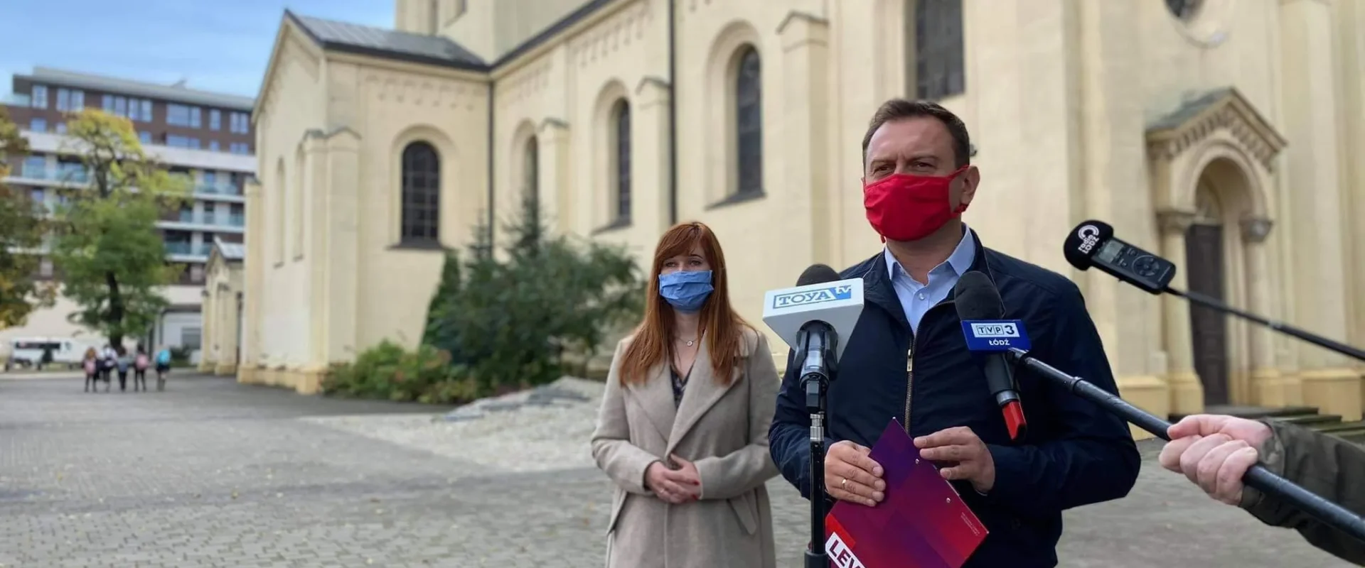 Dwóch dorosłych w maseczkach stoi przed zabytkowym kościołem i prowadzi konferencję prasową z mikrofonami, omawiając rolę MEN w niezastraszaniu dzieci i nauczycieli.