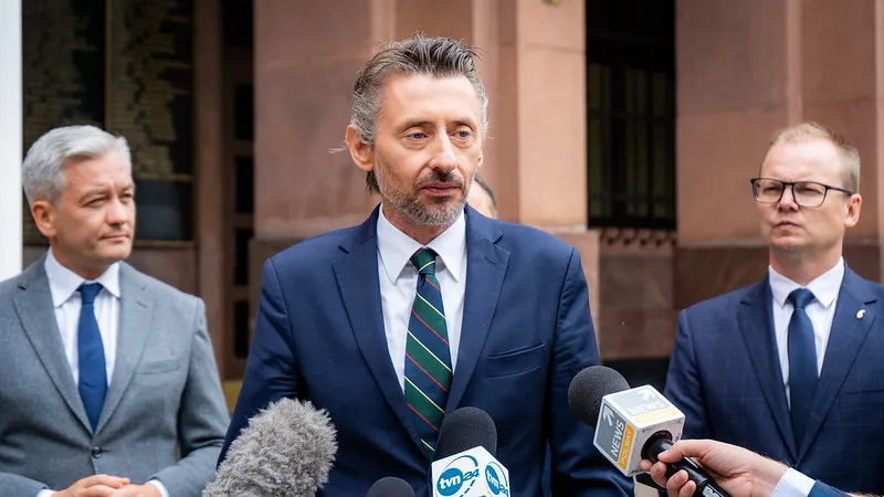 Trzech mężczyzn w garniturach stoi na konferencji prasowej; centralnie pozuje mężczyzna w niebieskim garniturze z zielono-paskanym krawatem, mówiący do mikrofonów, podczas gdy dwóch współpracowników stoi z boku.