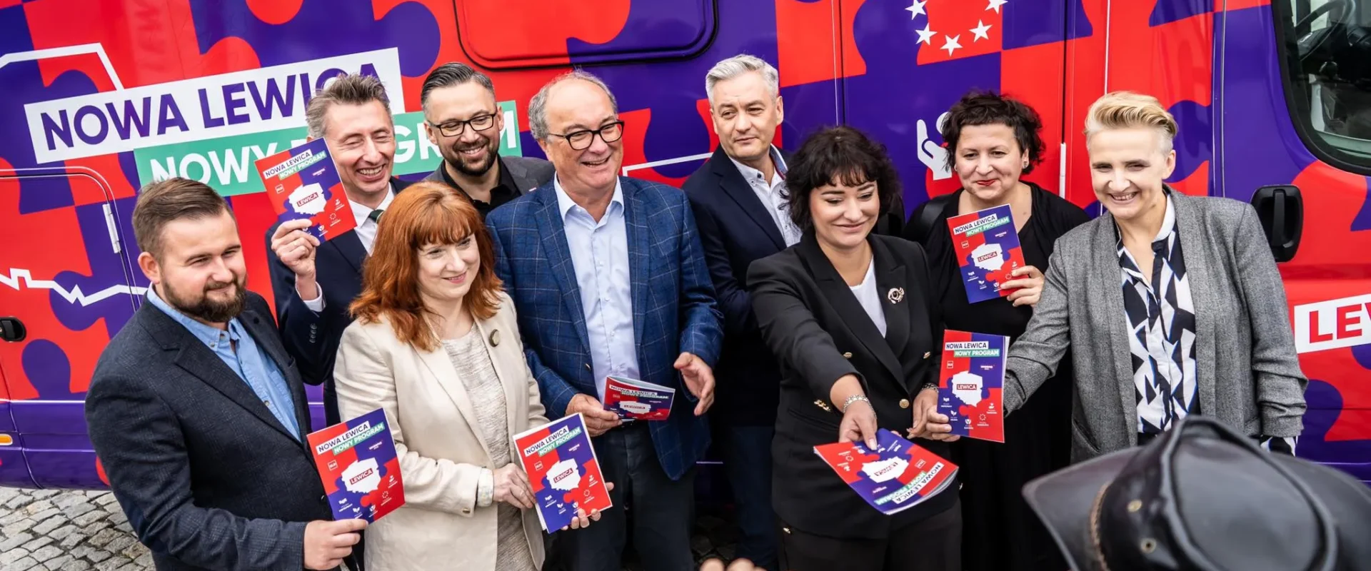 Grupa osób stojących obok kolorowego kampera kampanii Nowa Lewica, trzymających plakaty i broszury promujące konsultacje programowe.
