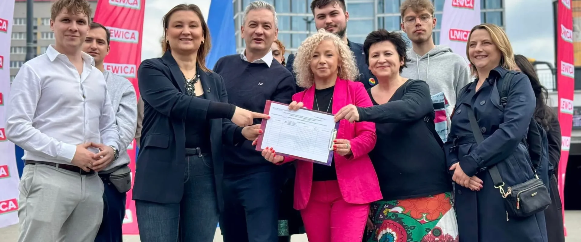Grupa osób na zewnątrz, trzymająca kartę z podpisami, stojąca w otoczeniu flag Lewicy przy nowoczesnym budynku, podczas zbierania podpisów pod rejestrację komitetu Lewicy do wyborów do Parlamentu Europejskiego.