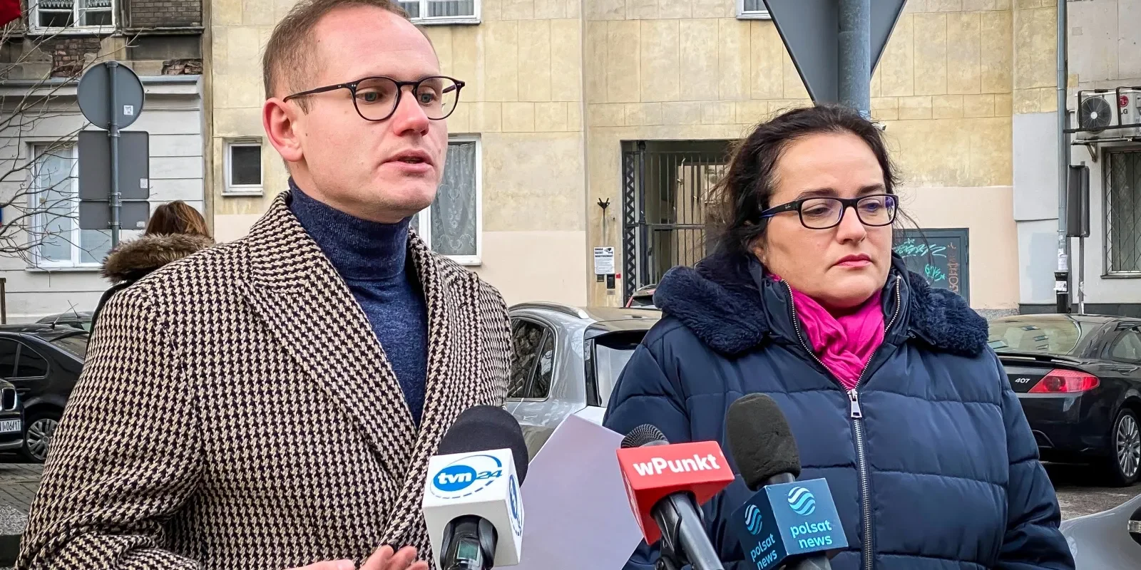 Dwóch rozmówców stojących na ulicy podczas konferencji prasowej; mężczyzna w beżowo‑czarnym pepitowym płaszczu i okularach oraz kobieta w granatowej kurtce z różowym szalem stoją obok mikrofonów z logotypami stacji telewizyjnych.