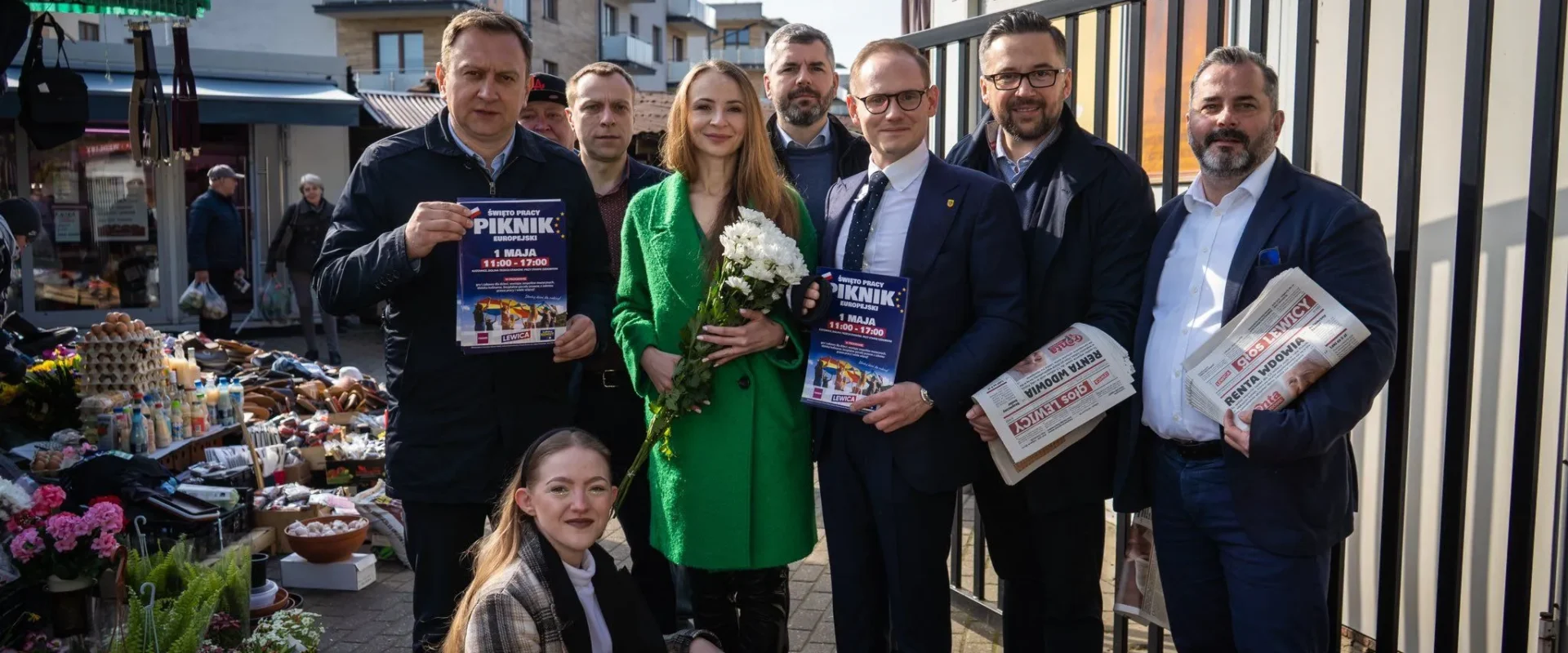 Na zdjęciu grupa osób trzymająca plakaty zapraszające na piknik z okazji Święta Pracy, stojąca na tle handlowym w słoneczny dzień.