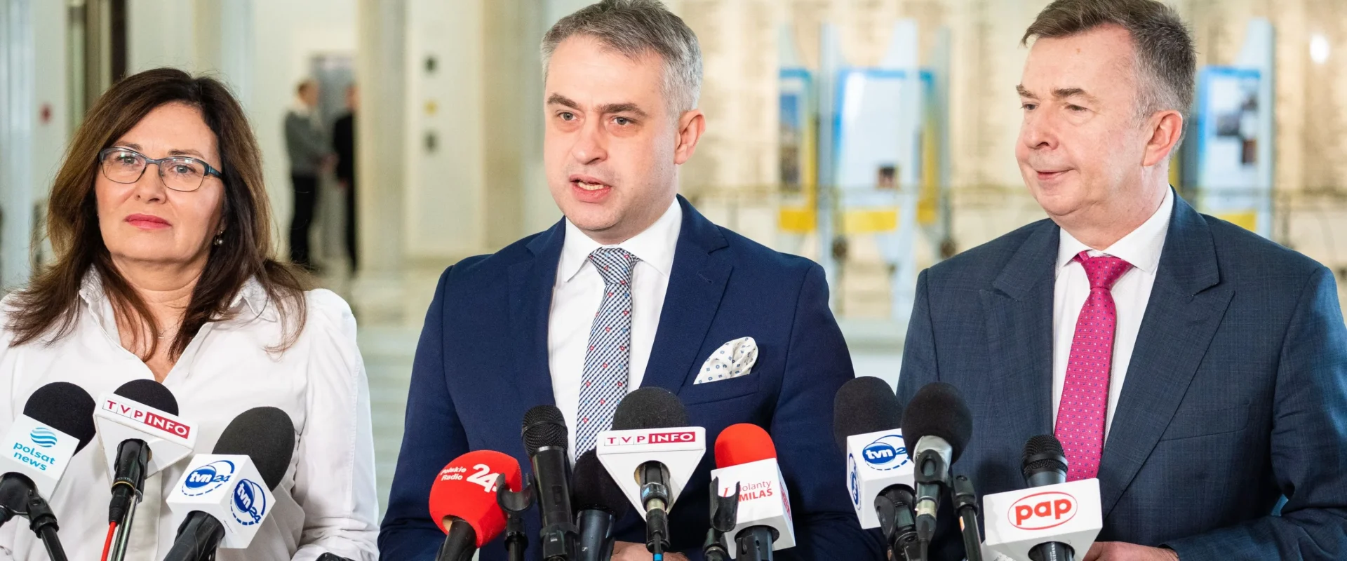 Na zdjęciu widoczni są trzej mówcy stojący przed mikrofonami podczas konferencji prasowej.