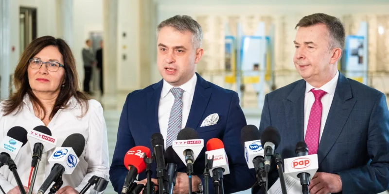 Na zdjęciu widoczni są trzej mówcy stojący przed mikrofonami podczas konferencji prasowej.