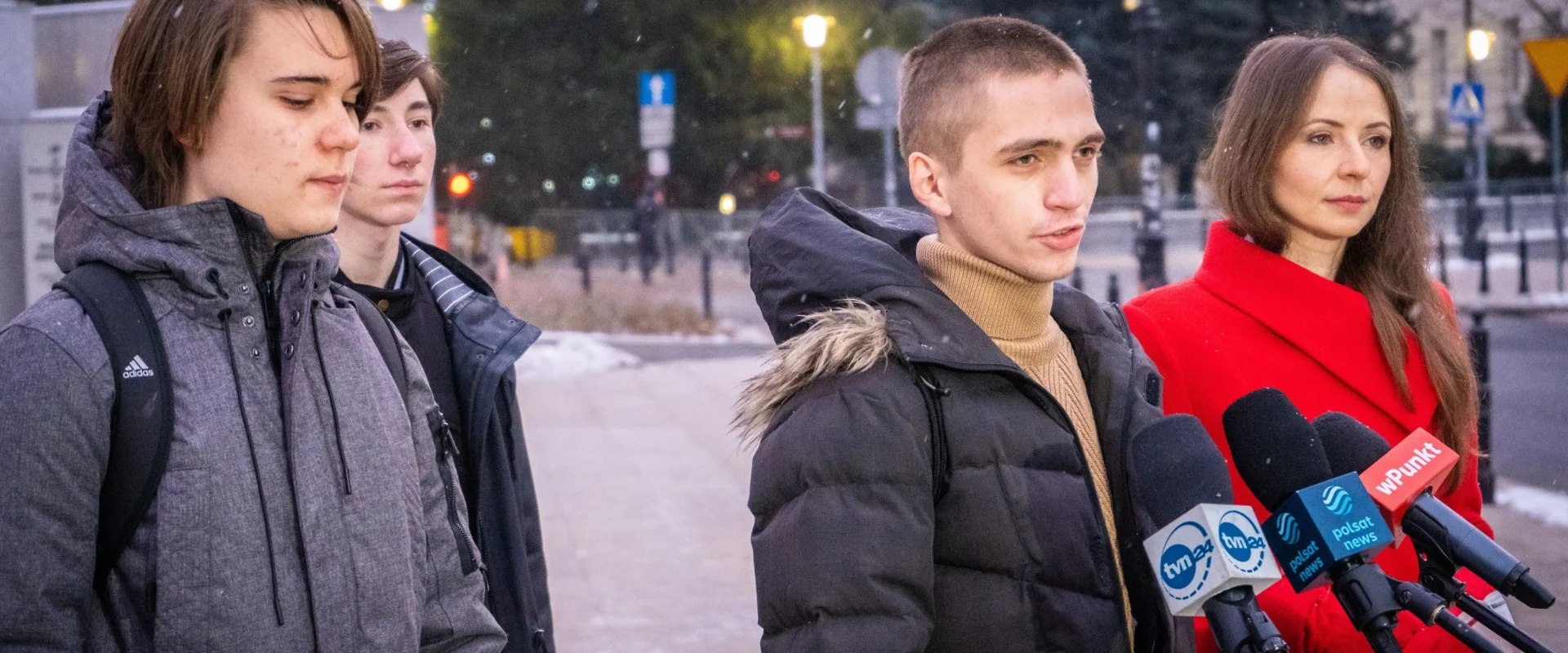 Grupa młodych ludzi i kobieta w czerwonym płaszczu stoi przed mikrofonami, podczas konferencji prasowej na zewnątrz w zimowej scenerii.
