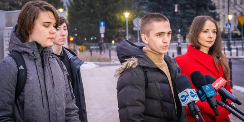 Grupa młodych ludzi i kobieta w czerwonym płaszczu stoi przed mikrofonami, podczas konferencji prasowej na zewnątrz w zimowej scenerii.