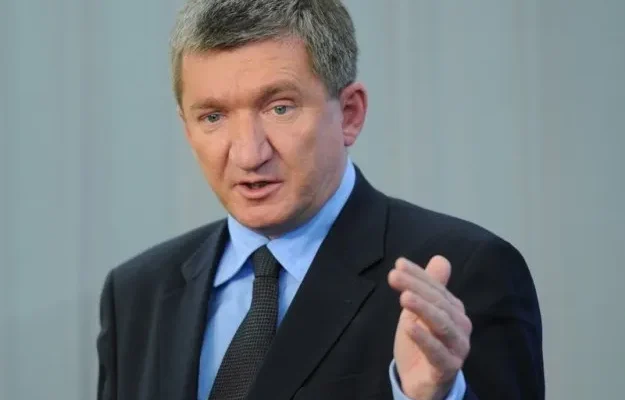 Mężczyzna w garniturze i krawacie przemawia, gestykulując jedną dłonią na jasnym tle.