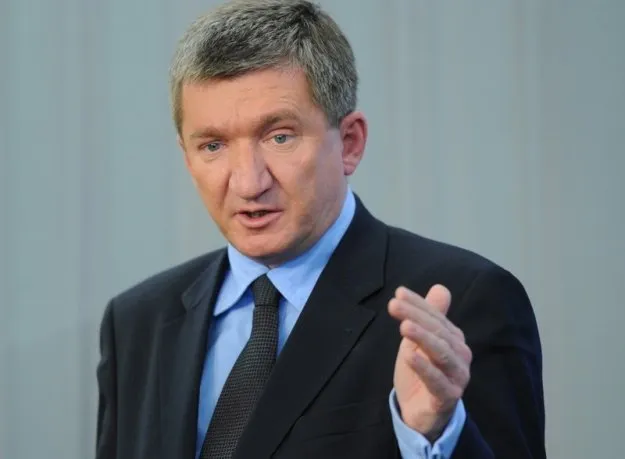 Mężczyzna w garniturze i krawacie przemawia, gestykulując jedną dłonią na jasnym tle.