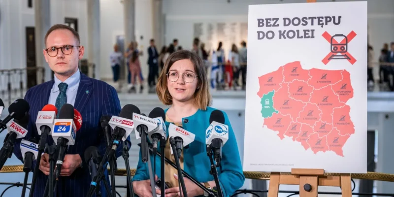 Dwójka mówców stoi przed mapą Polski z oznaczeniami, na której widnieje hasło "Bez dostępu do kolei", w czasie konferencji prasowej.