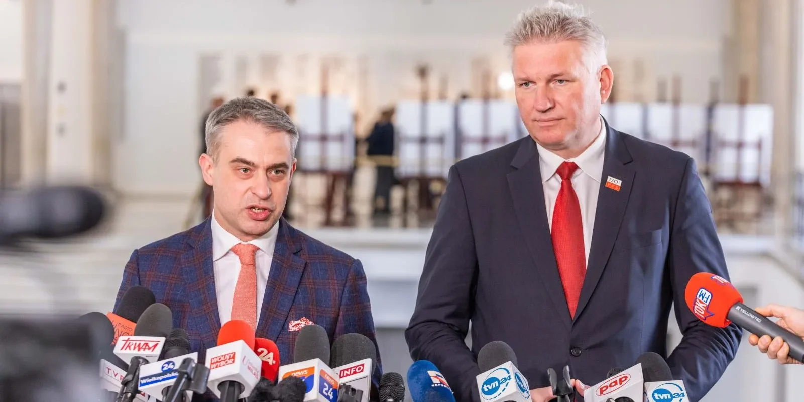 Dwaj mężczyźni w garniturach stoją przy mównicy otoczeni mikrofonami, podczas konferencji prasowej w jasnym wnętrzu.