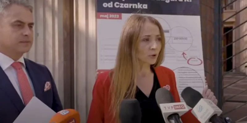 Kobieta w czerwonym żakiecie przemawia do mediów, stojąc obok mężczyzny w garniturze przed tablicą z wykresami.