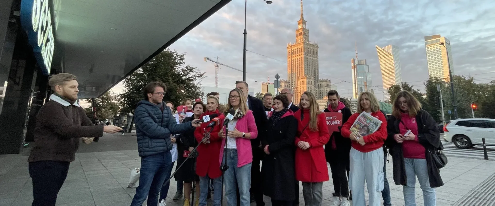 Grupa osób na placu przed nowoczesnym budynkiem prowadzi konferencję prasową z mikrofonami; w tle widać wysoki wieżowiec i inne budynki.