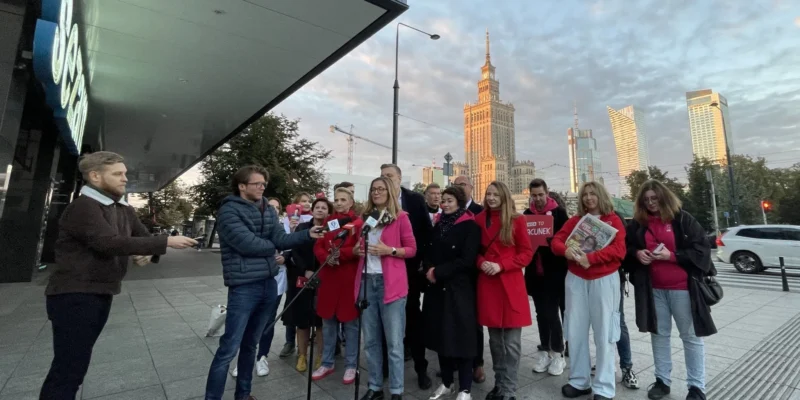 Grupa osób na placu przed nowoczesnym budynkiem prowadzi konferencję prasową z mikrofonami; w tle widać wysoki wieżowiec i inne budynki.