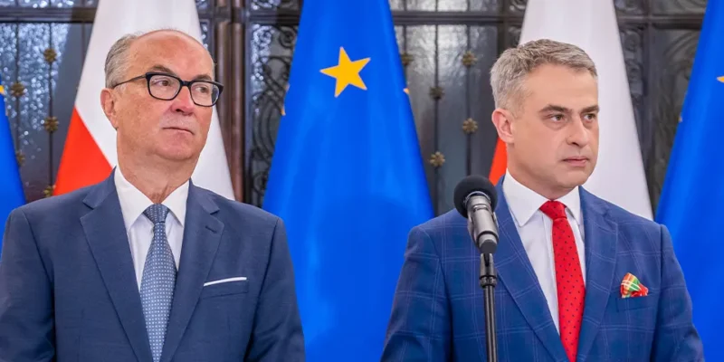 Dwie osoby w garniturach stojące przy mikrofonie podczas konferencji prasowej, z flagami Unii Europejskiej i Polski w tle.