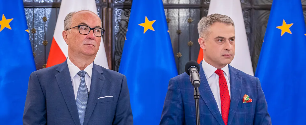 Dwie osoby w garniturach stojące przy mikrofonie podczas konferencji prasowej, z flagami Unii Europejskiej i Polski w tle.