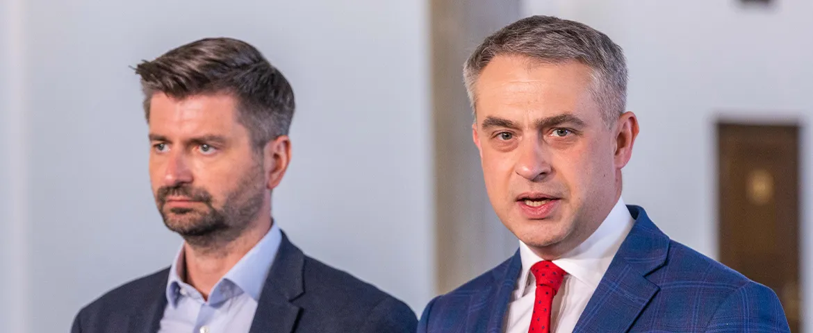 Dwóch mężczyzn w garniturach stoi w jasnym wnętrzu; po prawej stronie jeden z nich mówi, a po lewej słucha.