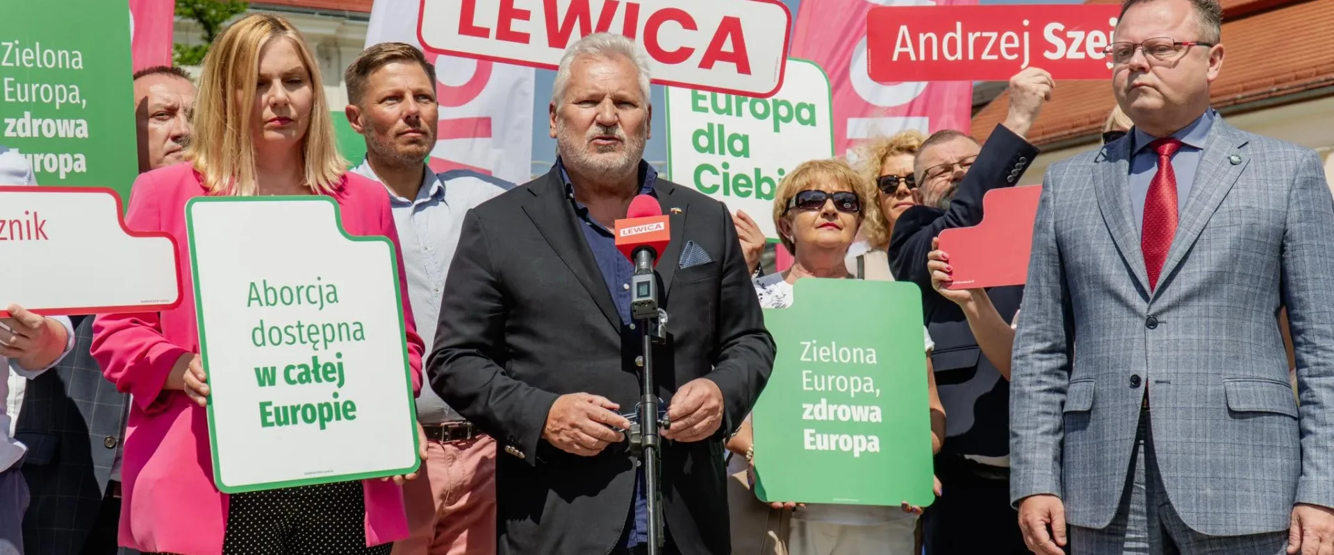 Grupa zwolenników Lewicy trzyma plakaty z hasłami „Zielona Europa, zdrowa Europa” i „Aborcja dostępna w całej Europie” podczas przemowy przy mikrofonie.